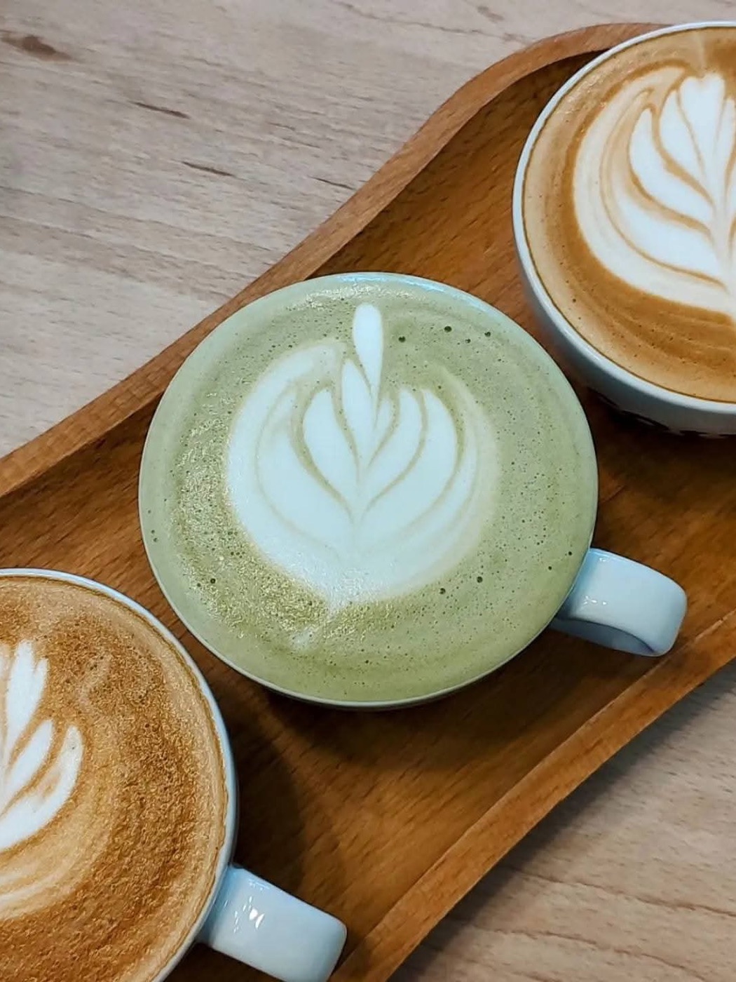 Matcha latte cu latte art, între două căni de cafea, pe o tavă de lemn